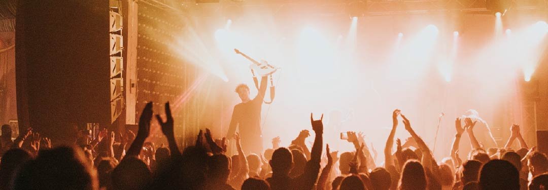 Energetic live music concert with a guitarist performing on stage under bright lights as a cheering crowd raises their hands.