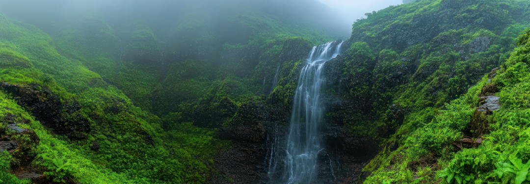 Lush green mountain valley with a cascading waterfall, surrounded by dense forests and mist-covered hills, showcasing India's scenic adventure destinations.