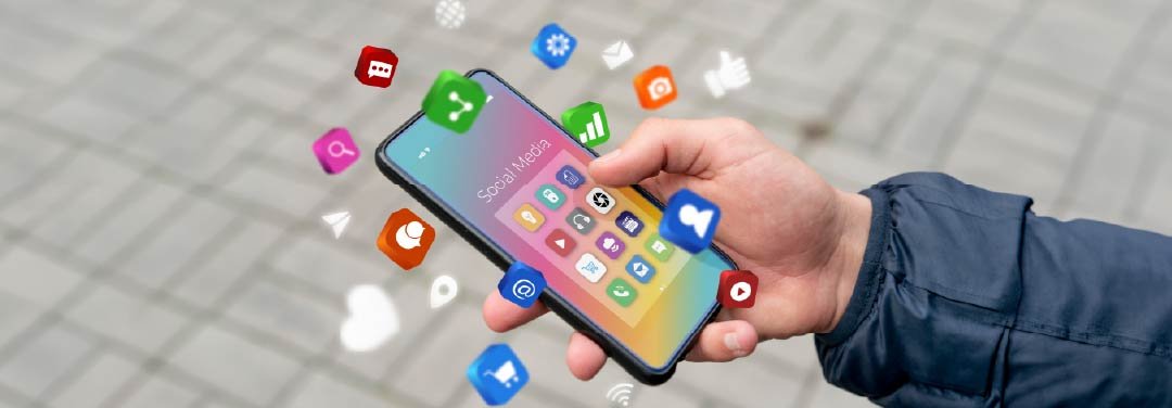 Person holding a smartphone displaying colourful social media app icons, with floating symbols representing messaging, notifications and digital connectivity.