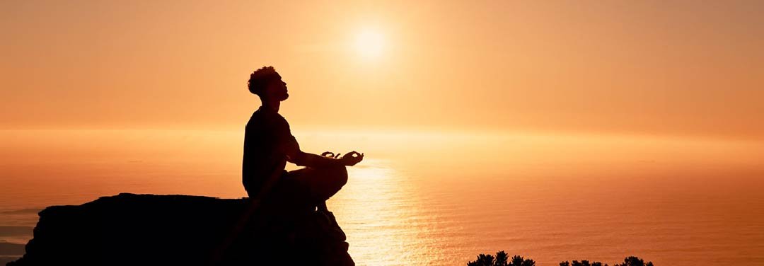 Silhouette of a person meditating on a rocky cliff overlooking the ocean at sunset.