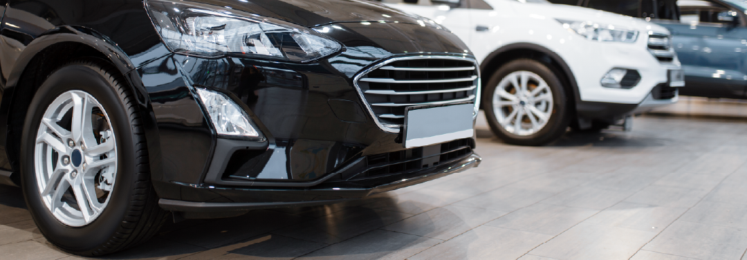 Close-up of a new black car displayed in an automobile showroom, with other vehicles in the background.