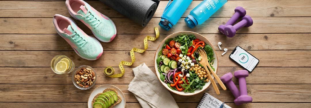 Healthy salad, running shoes, yoga mat, dumbbells, water bottles and fitness accessories arranged on a wooden floor.