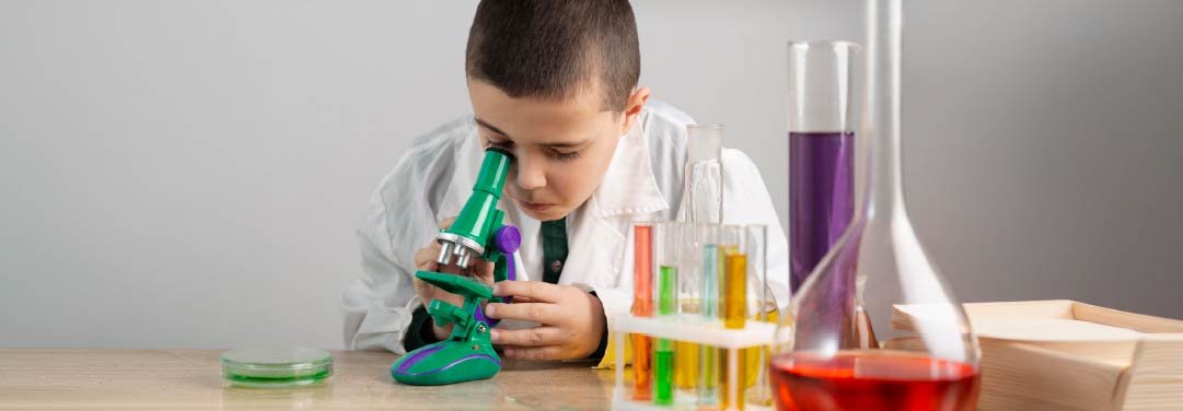Young boy wearing a white lab coat looking through a green microscope during a science experiment, with colourful liquids in test tubes, laboratory flasks and a petri dish arranged on a table.