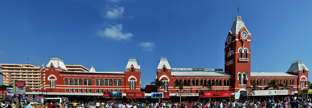 Historic Chennai Central railway station building with its distinctive red-brick architecture, clock tower and arched windows under a blue sky.