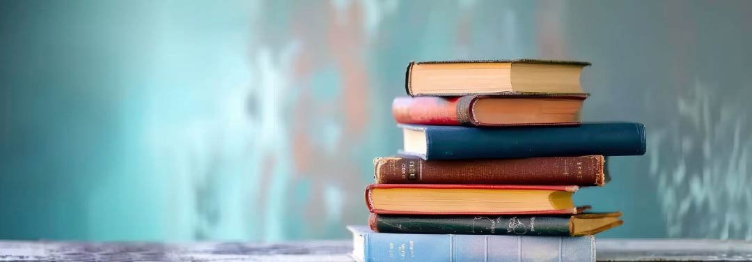 Stack of colourful hardcover books arranged on a wooden table against a soft blue background, representing South Asian literature and reading.