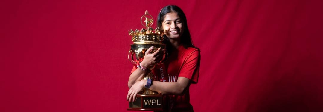 Indian cricketer Shreyanka Patil smiling while holding the Women's Premier League (WPL) trophy against a red background.