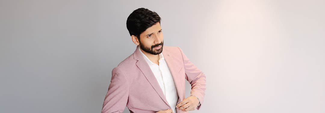 Editorial portrait of a bearded man wearing a pastel pink blazer and white shirt, posing against a minimalist light grey background.